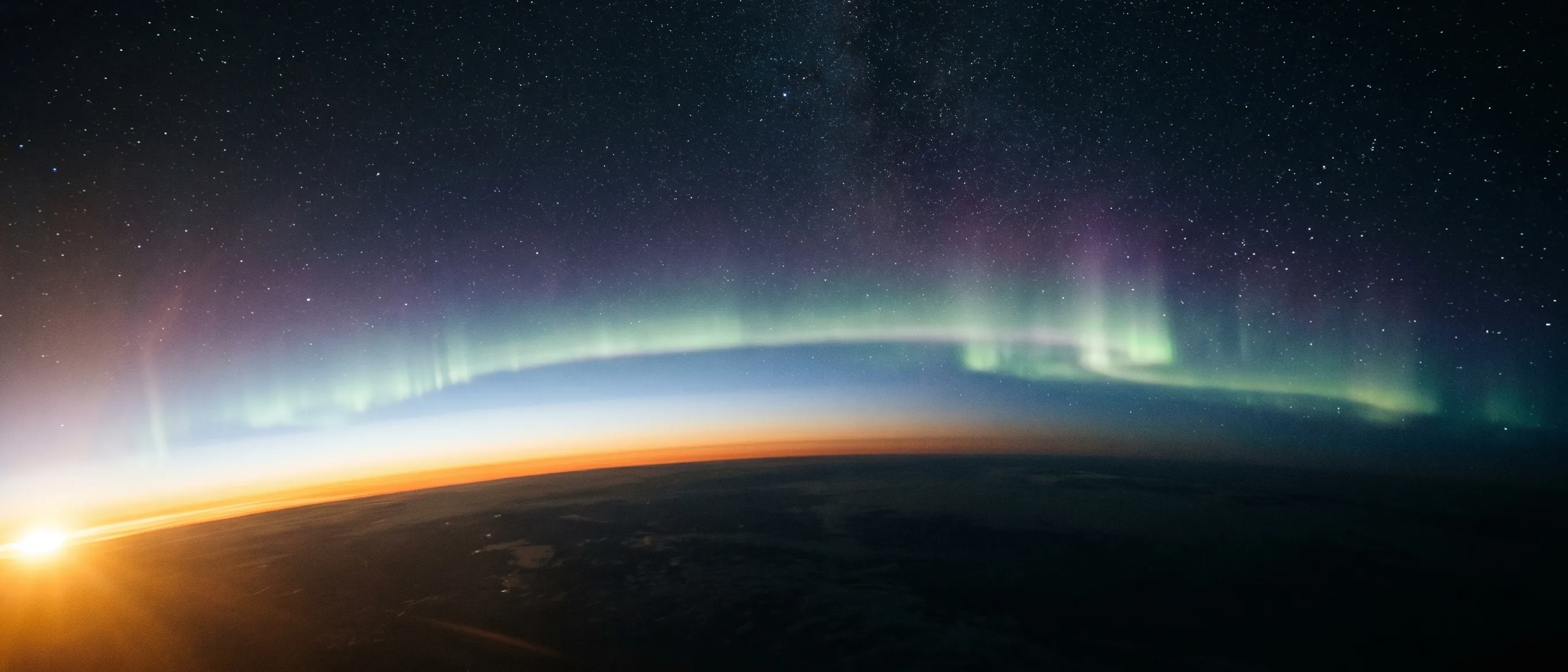 The curved edge of Earth glowing with a green-and-violet aurora beneath a star-filled sky, sunrise breaking gold at the horizon, seen from low orbit.