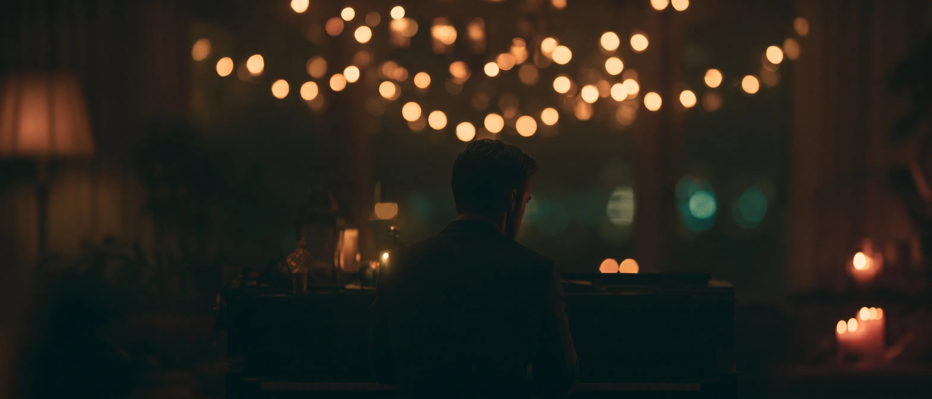 A man seen from behind, silhouetted at a piano in a candlelit room under a drift of warm string-light bokeh.