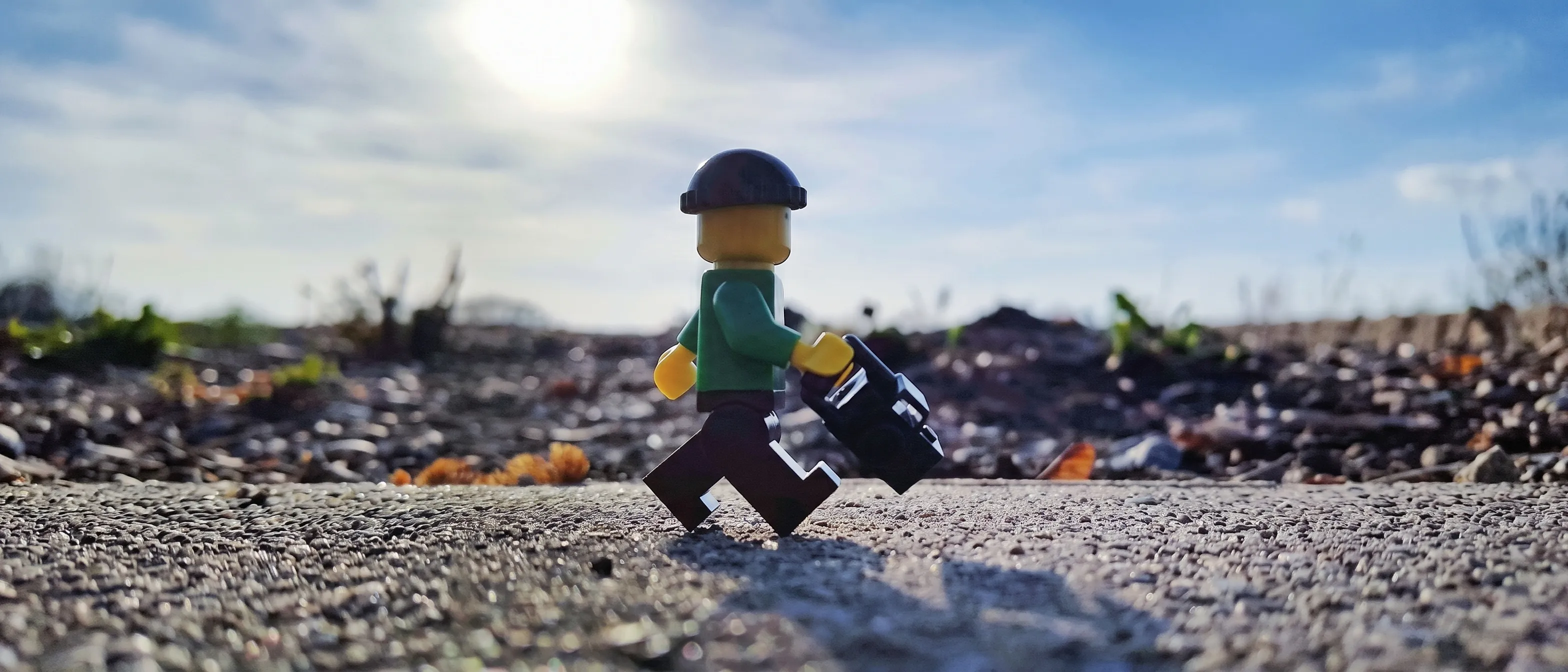 A green-jacketed Lego figure carrying a camera walks alone across a sunlit gravel lot toward a hazy, overexposed horizon.